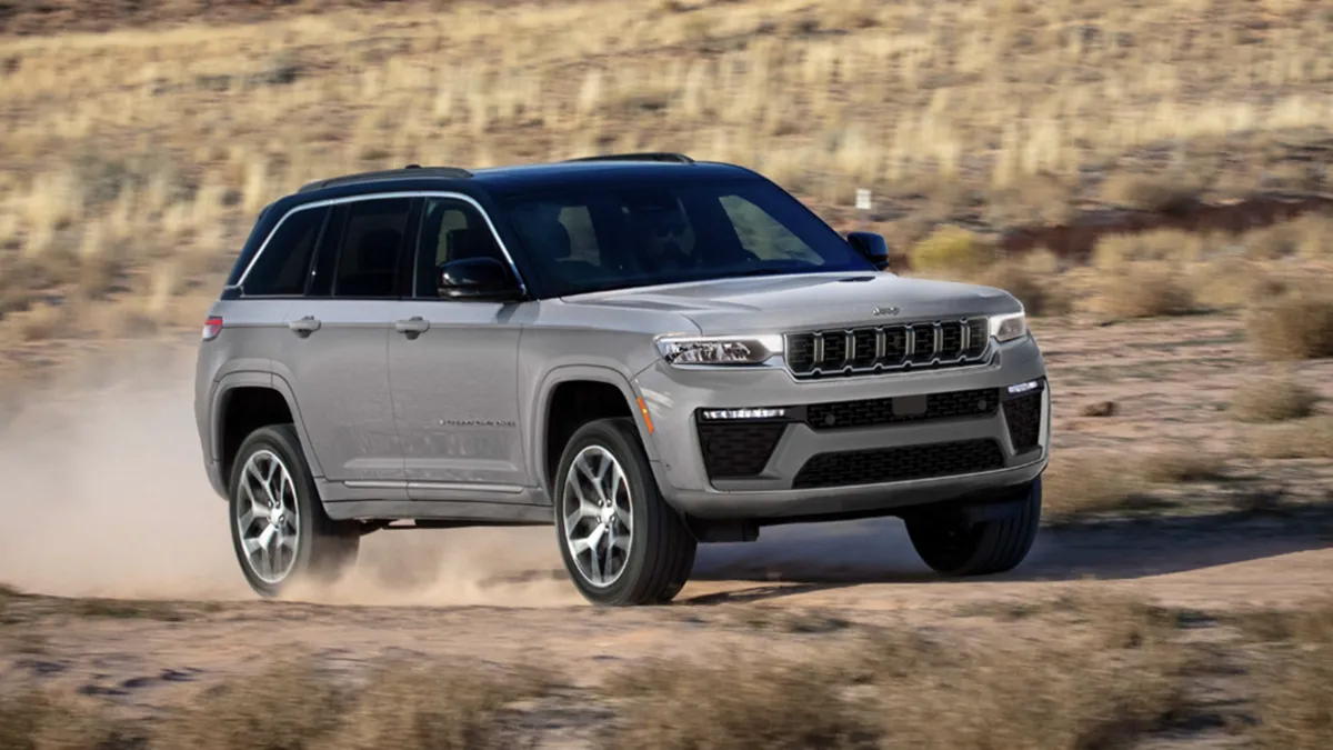 Front three-quarter view of a gray 2026 Jeep Grand Cherokee driving on a dusty road in dry terrain.