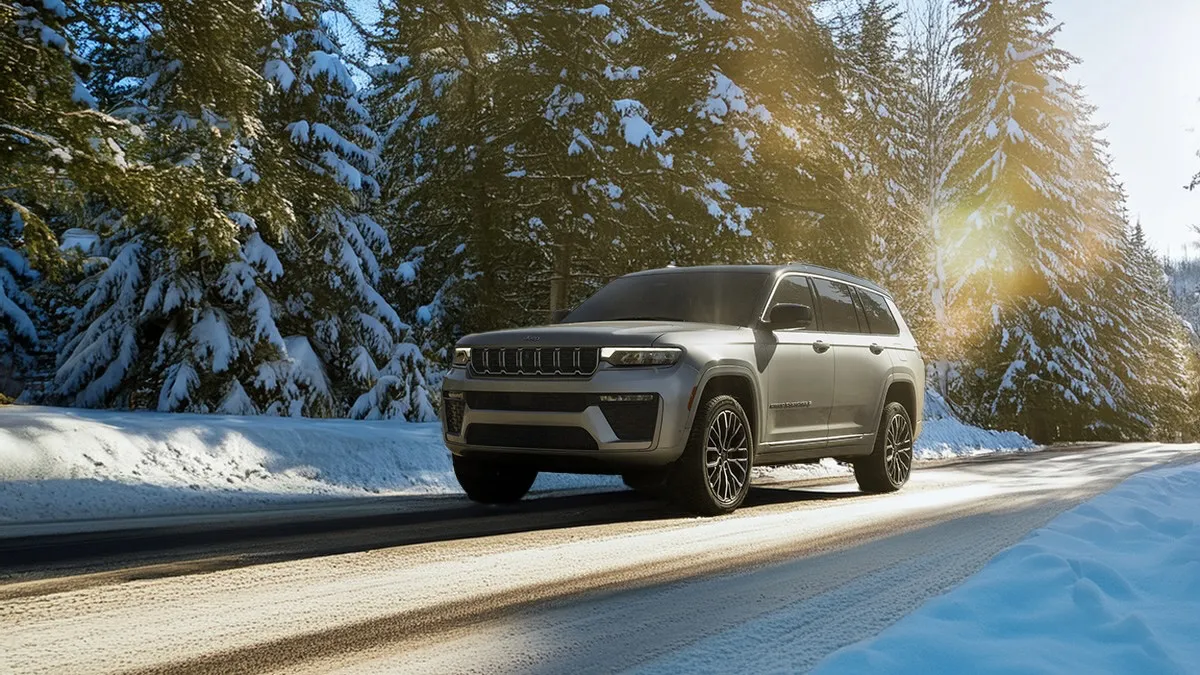 Front view of a black 2026 Jeep Grand Cherokee driving on a snowy road lined with trees.
