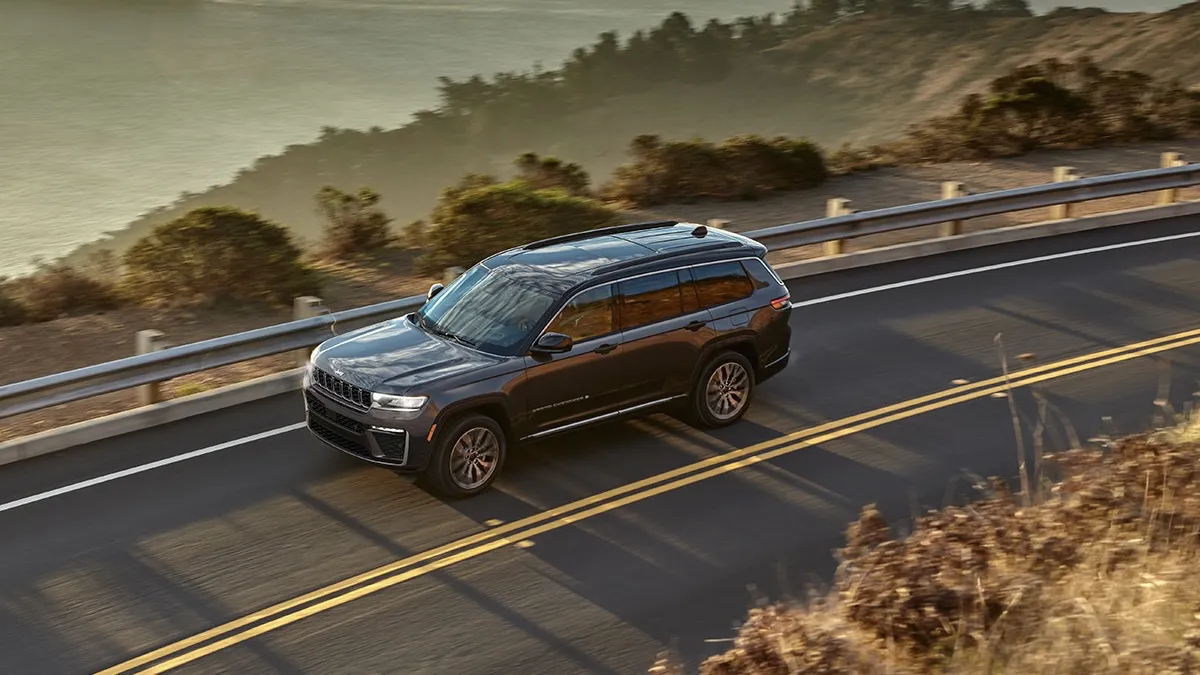 Front three-quarter view of a brown 2026 Jeep Grand Cherokee driving on a mountain road with a guardrail and curves.
