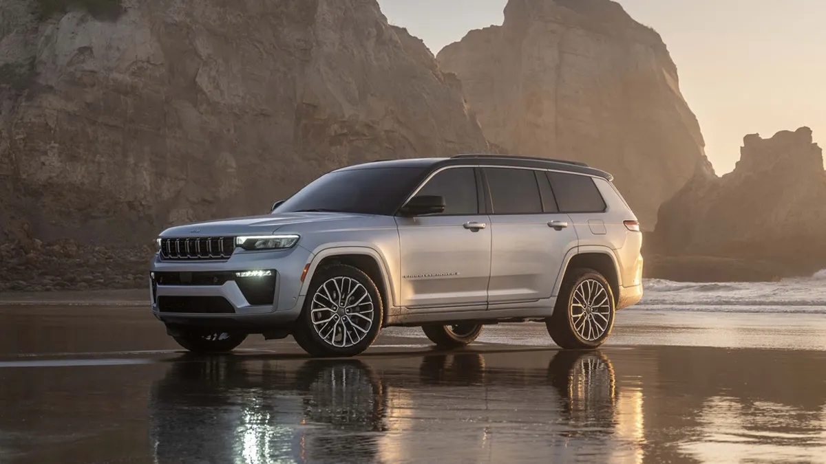 Front three-quarter view of a gray 2026 Jeep Grand Cherokee parked on a beach with rocky cliffs in the background.