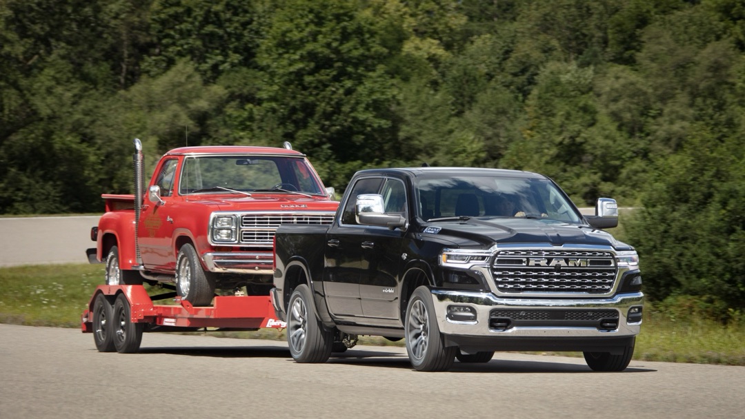Black 2026 Ram 1500 towing a trailer with a red pickup truck, driving on a road lined with trees.
