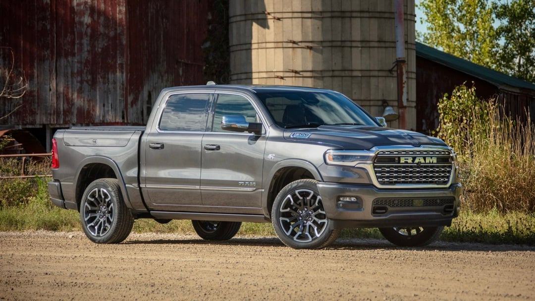 Vue de profil du Ram 1500 2026 gris stationné devant un bâtiment industriel avec végétation en arrière-plan.
			