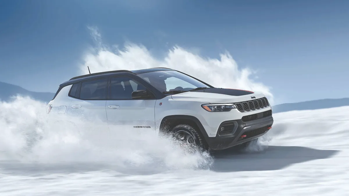 White 2026 Jeep Compass driving through the snow with a cloud of powder around the vehicle.

