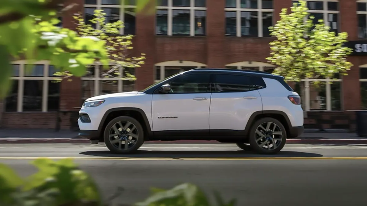 Side profile view of a white 2026 Jeep Compass driving in the city with buildings and trees in the background.
