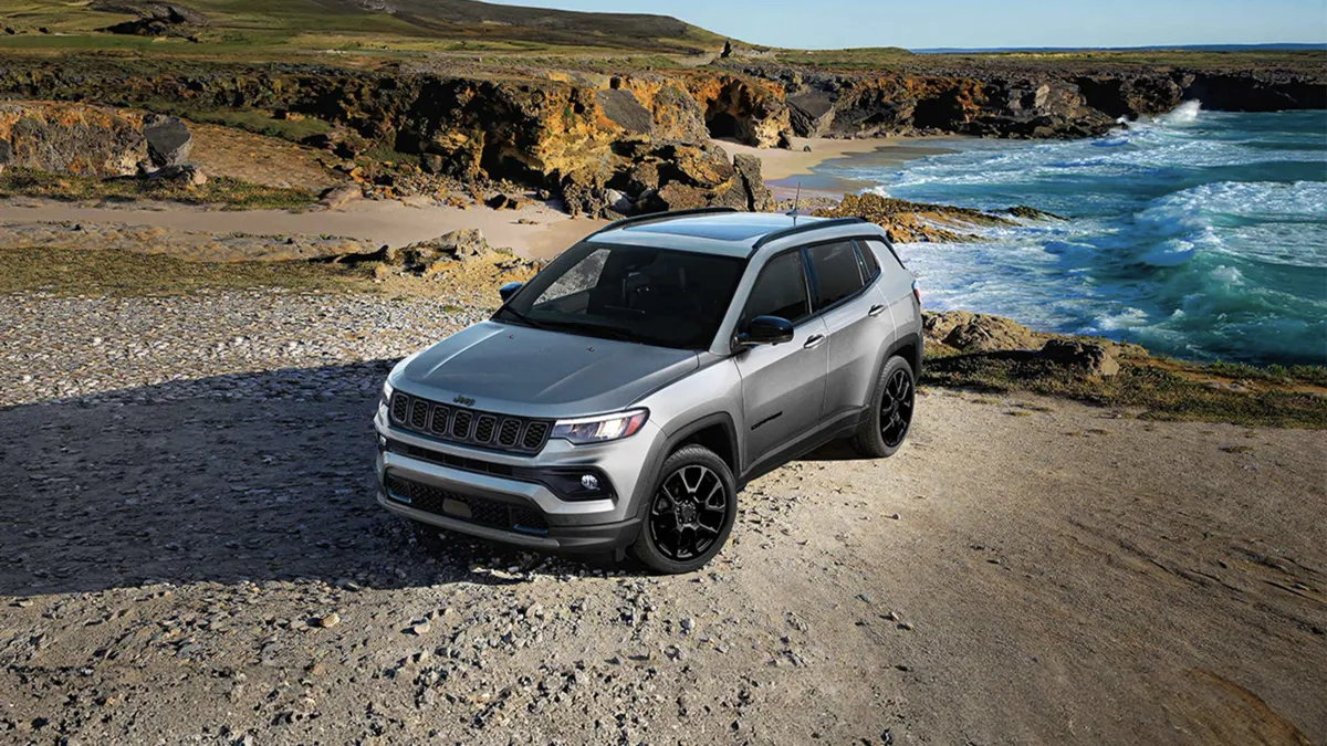 Front three-quarter view of a gray 2026 Jeep Compass parked by the sea with cliffs and shoreline in the background.

